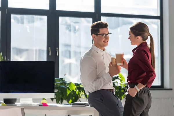 Giovani colleghi attraenti con tazze di carta di caffè parlare e flirtare in ufficio — Foto stock