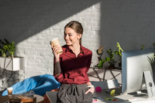 Sorridente jovem empresária com xícara de papel de café inclinando-se para trás na mesa de trabalho — Fotografia de Stock