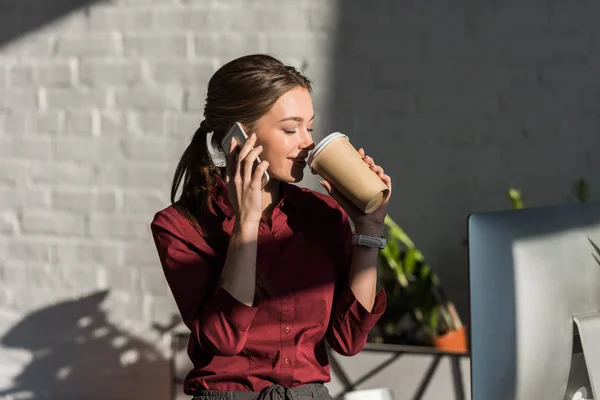 Hermosa joven empresaria beber café de la taza de papel y hablar por teléfono en la oficina - foto de stock