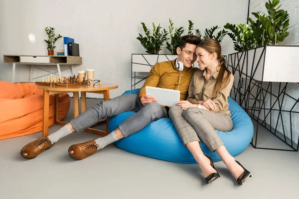 Pareja joven abrazándose usando tableta juntos mientras están sentados en la bolsa de frijoles - foto de stock