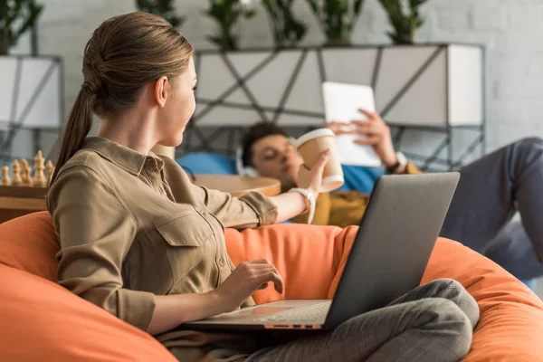 Jovem mulher bebendo café e trabalhando com laptop enquanto sentado em saco de feijão e passando xícara para namorado — Fotografia de Stock