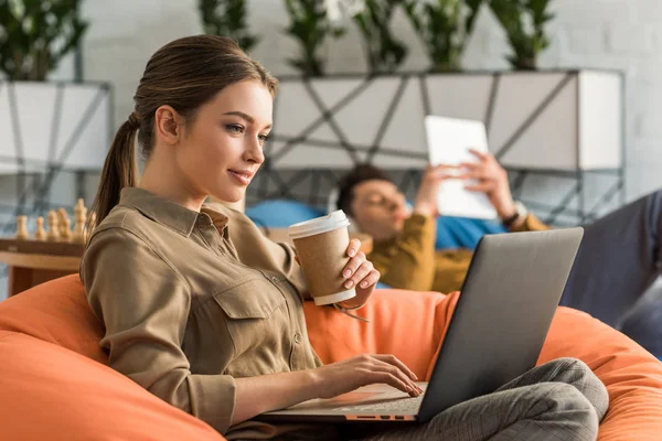 Молодая женщина пьет кофе и работает с ноутбуком, сидя в бобовом мешке — стоковое фото