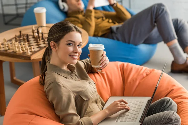 Щаслива молода жінка п'є каву і працює з ноутбуком, сидячи в бобовій сумці — стокове фото
