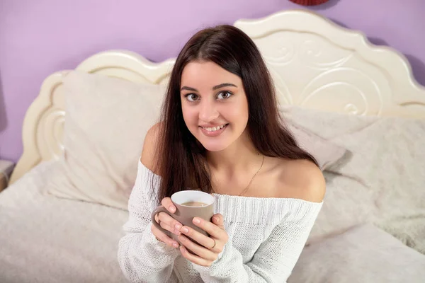 Brunette-jenta i seng med en kopp. En ung, smilende kvinne som ser på kamera og holder krus med kaffe. . – stockfoto