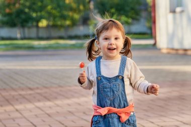 Yaz parkta şeker yemek küçük güzel bir kız. Denim tulum gülümseyen sevimli küçük kız. 2 yaşında bir çocuk portresi, yumuşak odak.