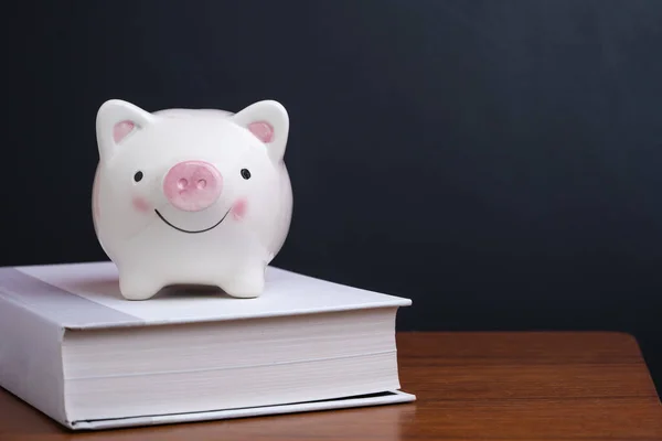 Financial knowledge, saving and investing education or scholarship  concept, pink smiling piggy bank on textbook on wooden table with dark blackboard or chalkboard background.