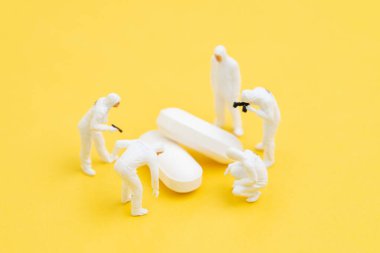 Koruyucu giysili minyatür doktorlar ve maske araştırmaları sarı arka planda beyaz haplar, Corona virüsü, COVID-19 grip ilacı veya virüs aşısı tıbbi araştırma konsepti.