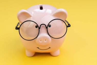 Sarı arka planda gözlük takan kumbara tasarruf, yatırım, bütçe ve finansal konsept olarak kullanılıyor..