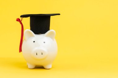 Sarı arka planda mezuniyet şapkası takan beyaz domuz bankası kopya alanı, eğitim fonu, burs veya üniversite ve üniversite konsepti için tasarruf.