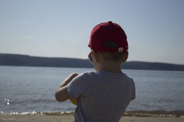 Küçük bir çocuk sahilde elinde topuyla su bakar.