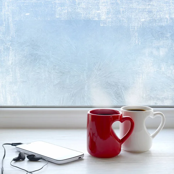 Duas xícaras de café, smartphone e fones de ouvido em um peitoril da janela. No fundo padrão gelado na janela como um fundo de Natal — Fotografia de Stock