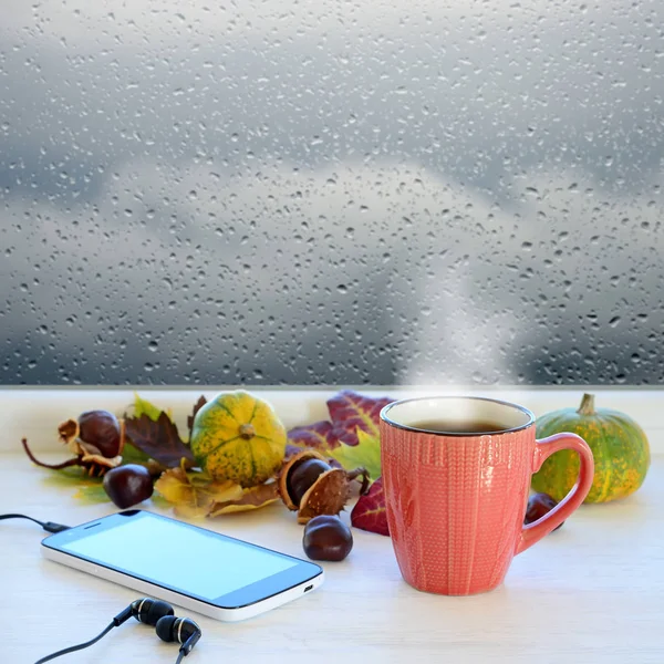一杯咖啡、 智能手机和耳机，秋天的落叶，南瓜和栗子在窗台上。在后台窗口与雨滴和云 — 图库照片