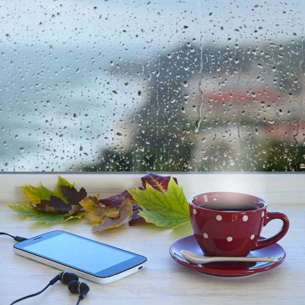 杯的咖啡、 智能手机、 耳机和秋天叶子在窗台上。在后台窗口与雨滴和云 — 图库照片