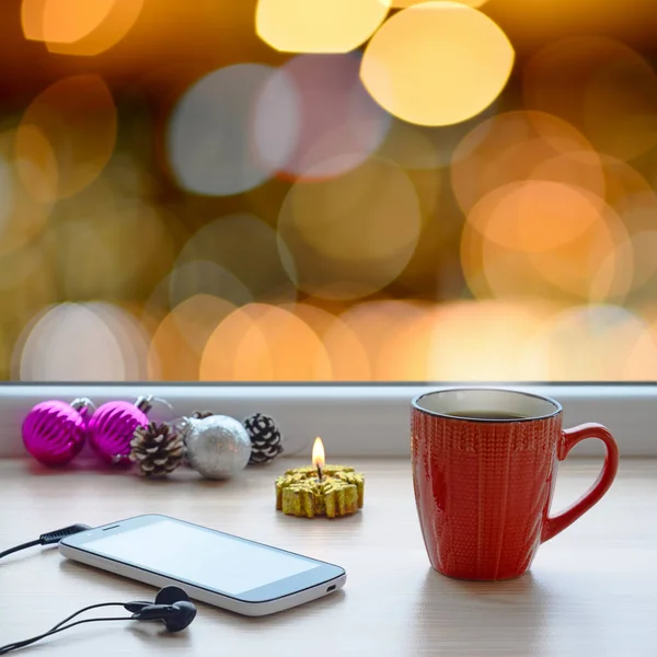 Xícara de café, smartphone, fones de ouvido e ornamentos de árvore de Natal em um peitoril da janela. No fundo, as luzes brilhantes da cidade — Fotografia de Stock