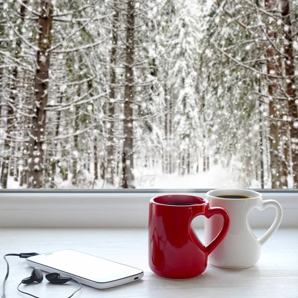 Two coffee cups, smartphone and headphones on a windowsill. In the background, a beautiful winter forest in snow — Stock Photo, Image
