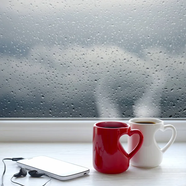 两杯咖啡、 智能手机和耳机在窗台上。在后台窗口与雨滴和云 — 图库照片