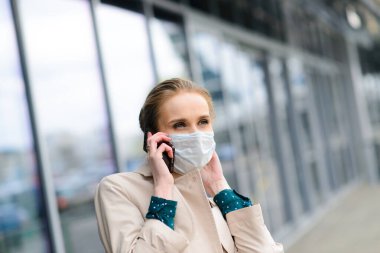 Maske takan ve şehirde telefonla konuşan genç bir iş kadını.