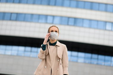 Maske takan ve şehirde telefonla konuşan genç bir iş kadını.