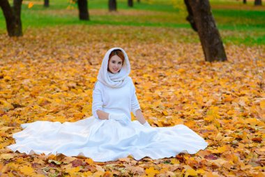 Uzun beyaz gelinlikli tatlı kız kırsal yolda poz veriyor ormandaki sonbahar ağaçları arasında altın saat atmosferinde.