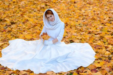 Uzun beyaz gelinlikli tatlı kız kırsal yolda poz veriyor ormandaki sonbahar ağaçları arasında altın saat atmosferinde.