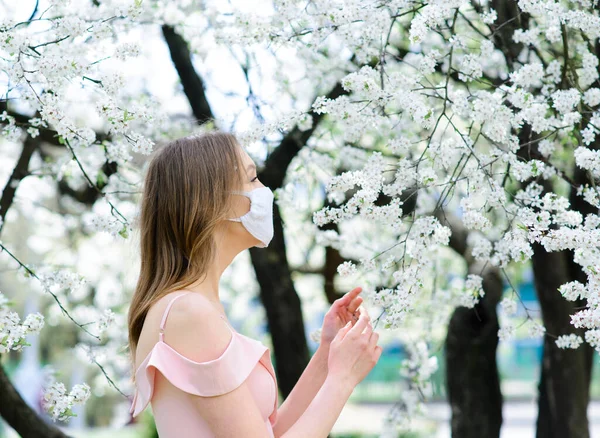 Közelkép egy érzékeny lányról fehér blúzban egy virágzó cseresznyefa alatt, a koronavírus maszkjával.. — Stock Fotó