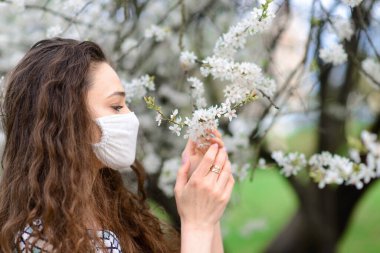 Coronavirus 'un çiçekli maskesiyle çiçek açan kiraz ağacının altında beyaz bir bluz içindeki hassas kızın portresini kapat..