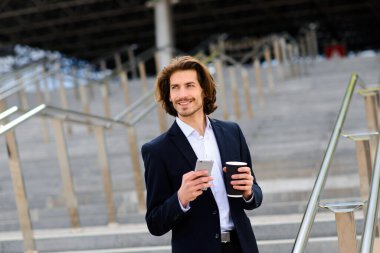 Tam takım elbiseli modern kendine güvenen genç işadamı arka planda şehir manzarasıyla dışarıda duruyor.