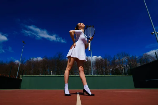 コートの上でテニスラケットを持って白いドレスとヒールでセクシーな女の子のテニス選手 若い女性はテニス スポーツをしている — ストック写真
