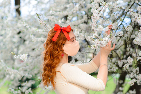 A beautiful red-haired young female in protective face mask stands near blossoming cherry tree during quarantine due to the coronavirus