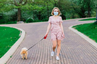 Çiçek açan bir ağacın arka planında köpeği olan mavi maskeli güzel bir kadın. Koronavirüs salgını.