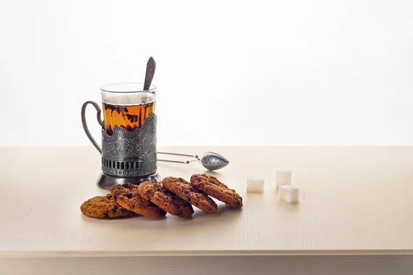 Tea in a Cup holder, on a light table-top under an oak, with oatmeal cookies, lump sugar, a teaspoon