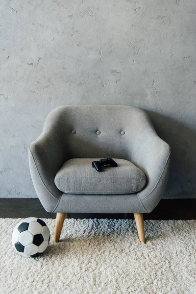 Soccer ball near grey armchair with gamepad — Stock Photo, Image