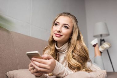 mutlu genç kadın evde kanepede Smartphone kullanarak ve uzağa arıyorsunuz