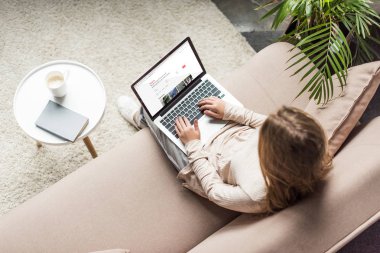 yüksek açılı görünüş kadının evde kanepede oturan ve airbnb Web sitesinde ekran ile dizüstü bilgisayar kullanarak