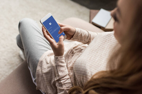 cropped shot of woman on couch using smartphone with facebook app on screen