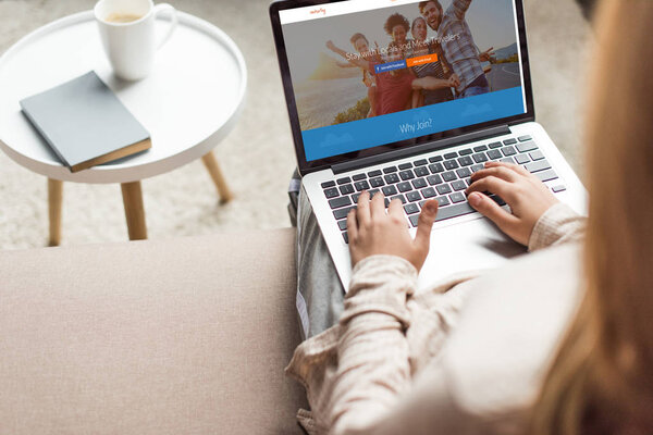 cropped shot of woman at home sitting on couch and using laptop with couchsurfing website homepage on screen