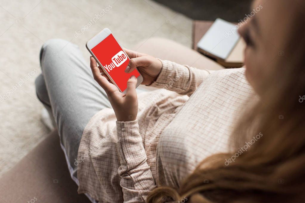 Cropped shot of woman on couch using smartphone with youtube logo on screen