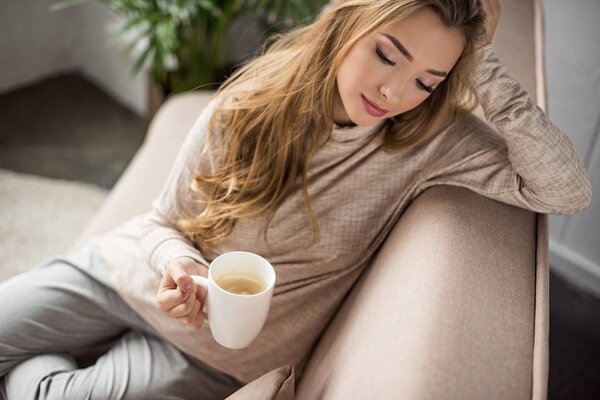 beautiful young woman in warm sweater drinking coffee on cozy couch