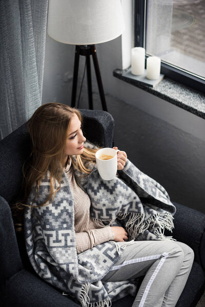 high angle view of beautiful young woman drinking coffee in cozy armchair with blanket