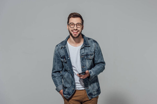 cheerful young man holding smartphone and smiling at camera isolated on grey