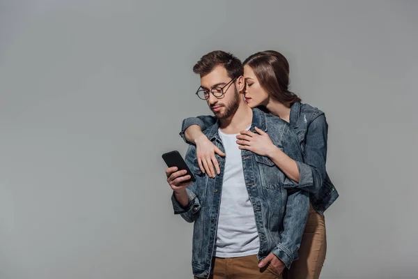 Jovem Mulher Abraçando Homem Bonito Óculos Segurando Smartphone Isolado Cinza — Fotografia de Stock