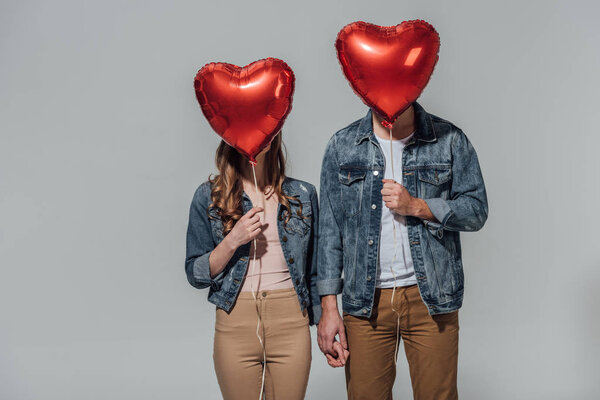 young couple hiding faces behind red heart shaped balloons isolated on grey