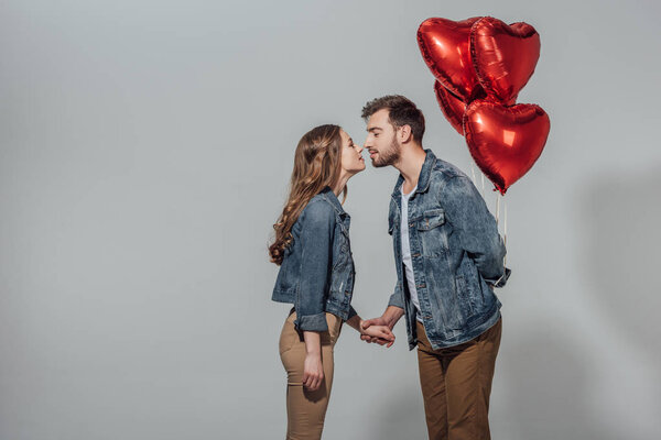 side view of young couple able to kiss while man holding red heart shaped balloons isolated on grey 