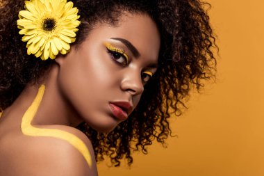 Young tender african american woman with artistic make-up and gerbera in hair looking at camera isolated on orange background clipart
