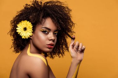 Young bright african american woman with artistic make-up and gerbera in hair touching her hair isolated on orange background clipart