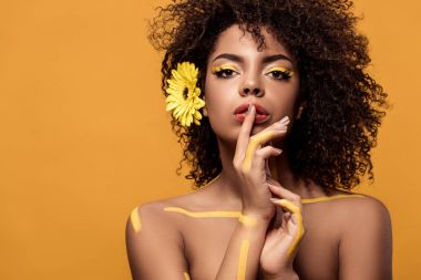 Şehvetli Afro-Amerikan olan kadın sanatsal makyaj ve saç üzerinde turuncu arka plan izole dudaklarında parmak tutan gerbera