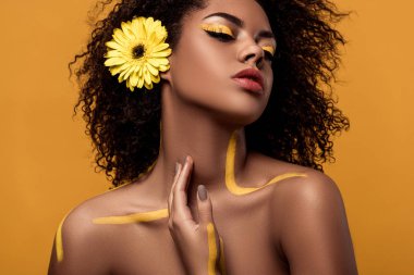 Stylish african american woman with artistic make-up and gerbera in hair tenderly touches her skin isolated on orange background clipart