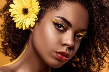 Yakın çekim sanatsal makyaj ve saç üzerinde turuncu arka plan izole gerbera genç şehvetli Afrikalı-Amerikalı kadın portresi
