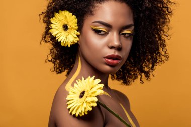 Young bright african american woman with artistic make-up and gerbera in hair looking away isolated on orange background clipart