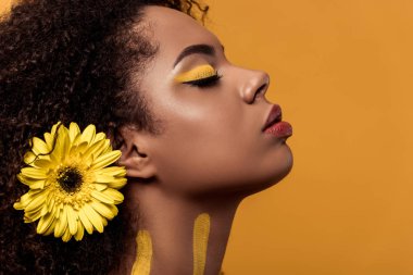 Stylish african american woman with artistic make-up and gerbera in hair dreaming isolated on orange background clipart